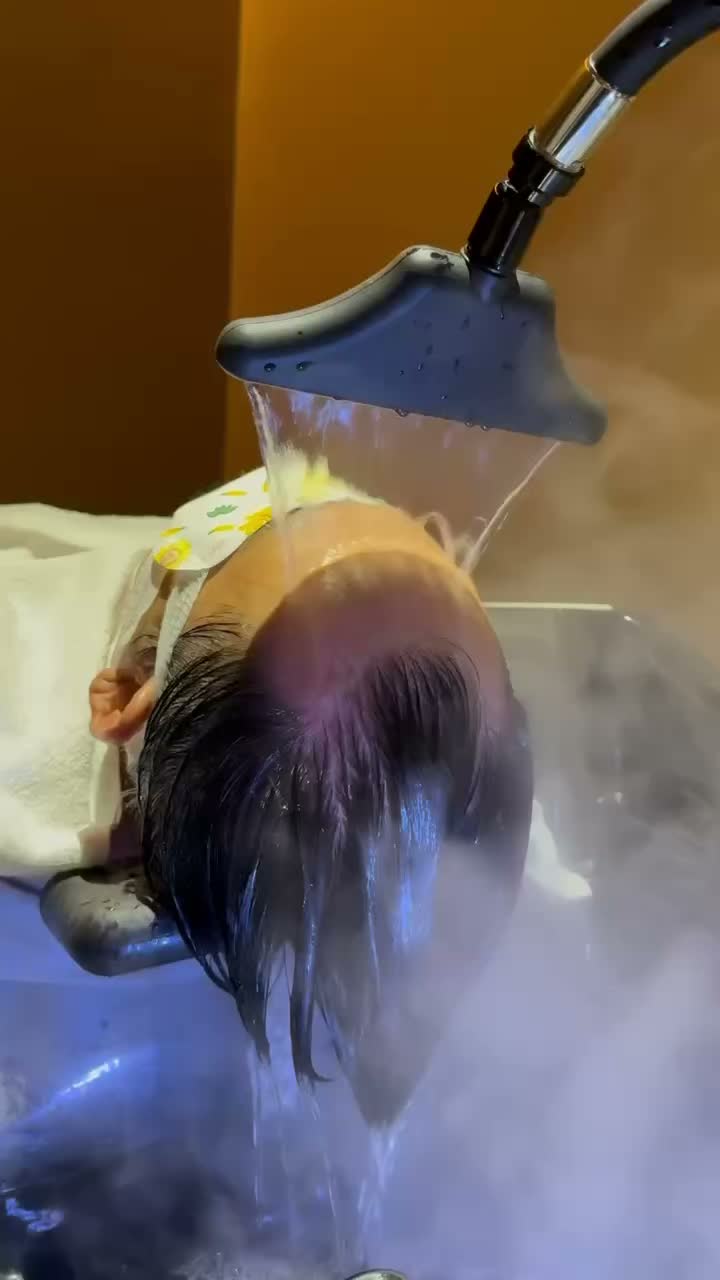 Hair wash treatment with water flowing over a client’s head under a salon rinse nozzle.